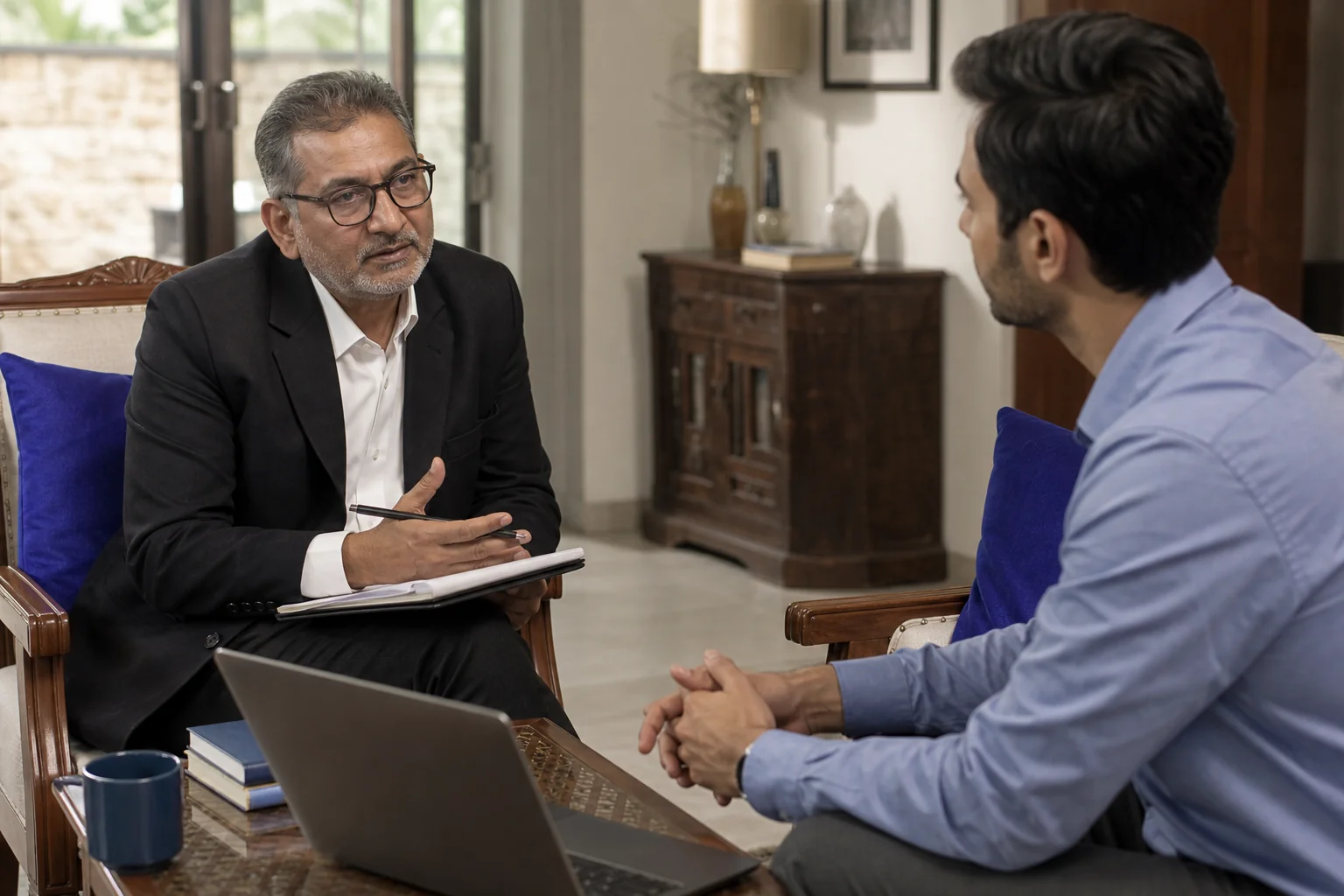 Photorealistic photo of a senior Indian male lawyer in his 50s wearing formal attire, sitting across a small table from a younger professional client in a warmly lit living room in Delhi, both engaged in a calm conversation, laptop and
