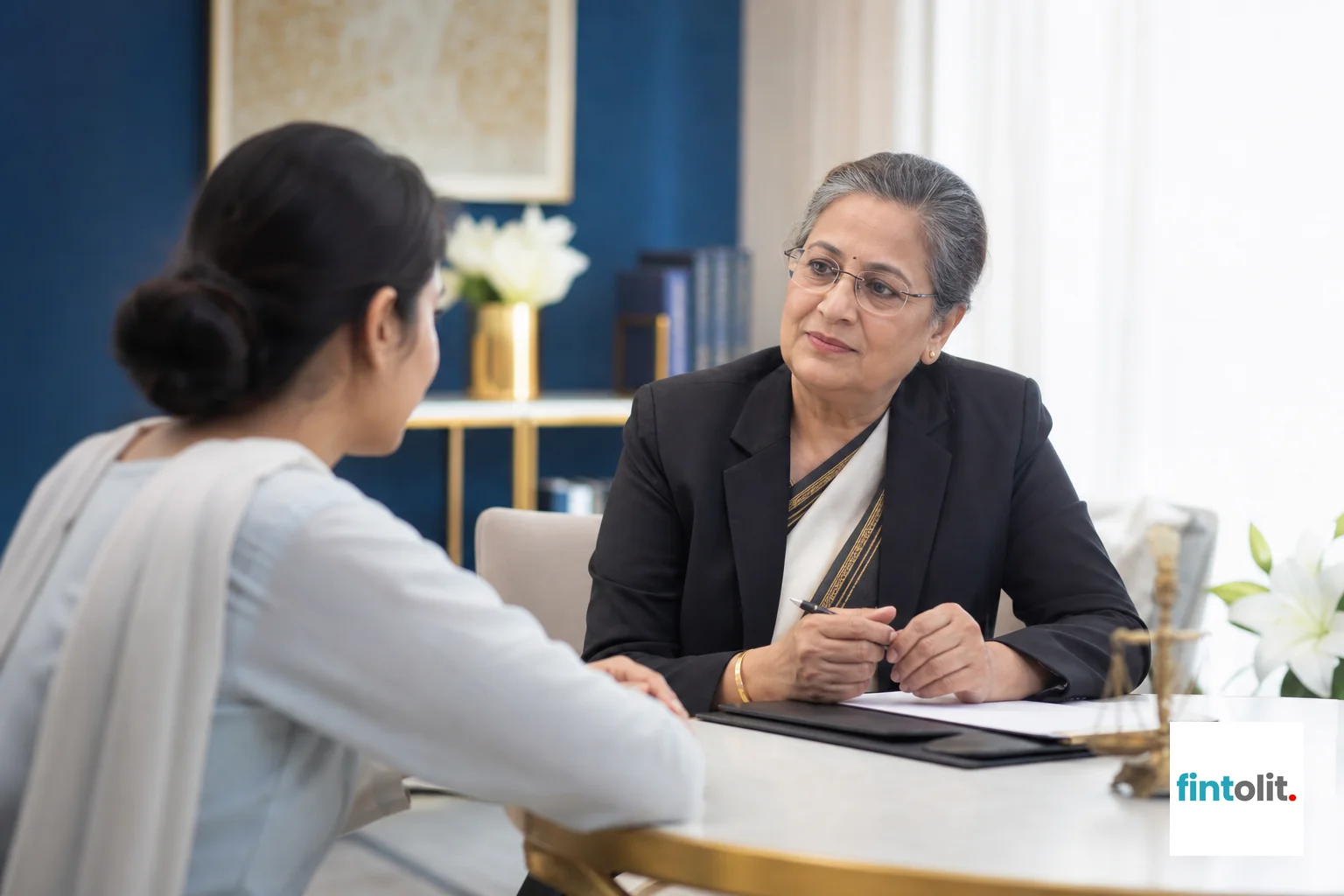 Photorealistic photo of a compassionate senior Indian female lawyer in professional attire talking with a woman client across a small table in a bright, modern consultation room, both engaged in calm conversation, soft daylight, indigo and